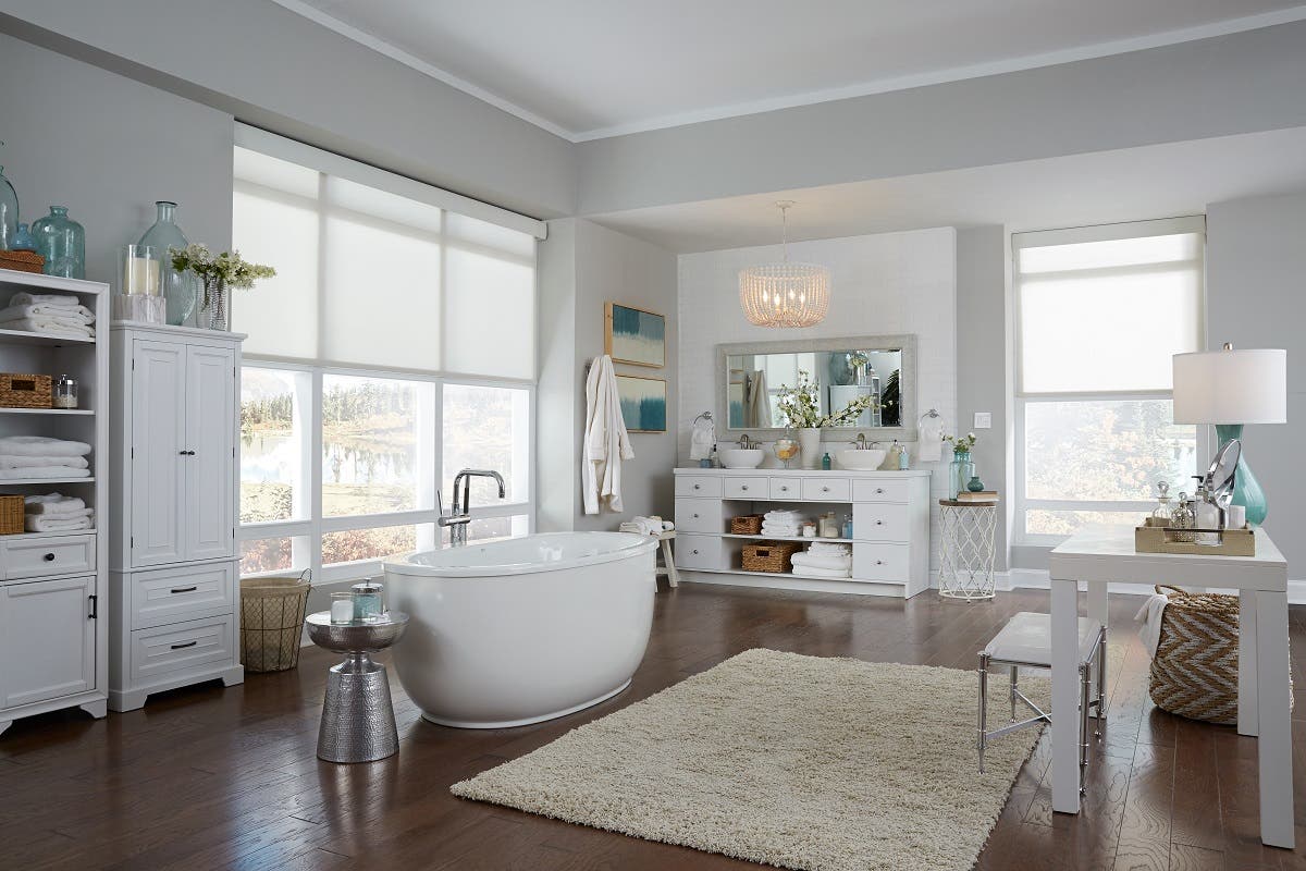 Spacious white bathroom with soaking tub in the middle and partially open white sheer roller shades